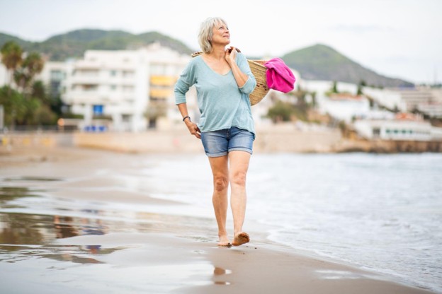A woman walking confidently along a beach wearing jean shorts and a loose pastel top.
